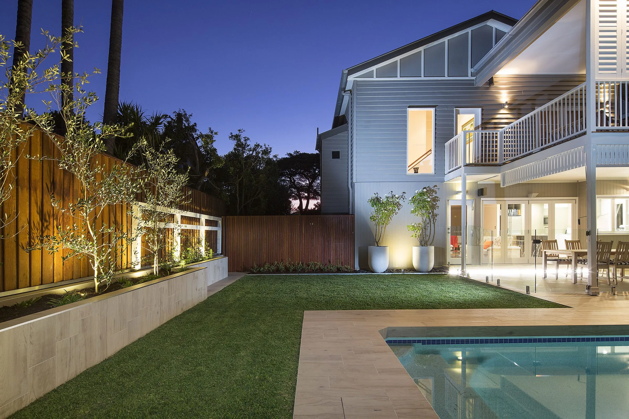 Twilight garden with a row of standard olive trees, timber batten fencing, and a frameless glass pool fence