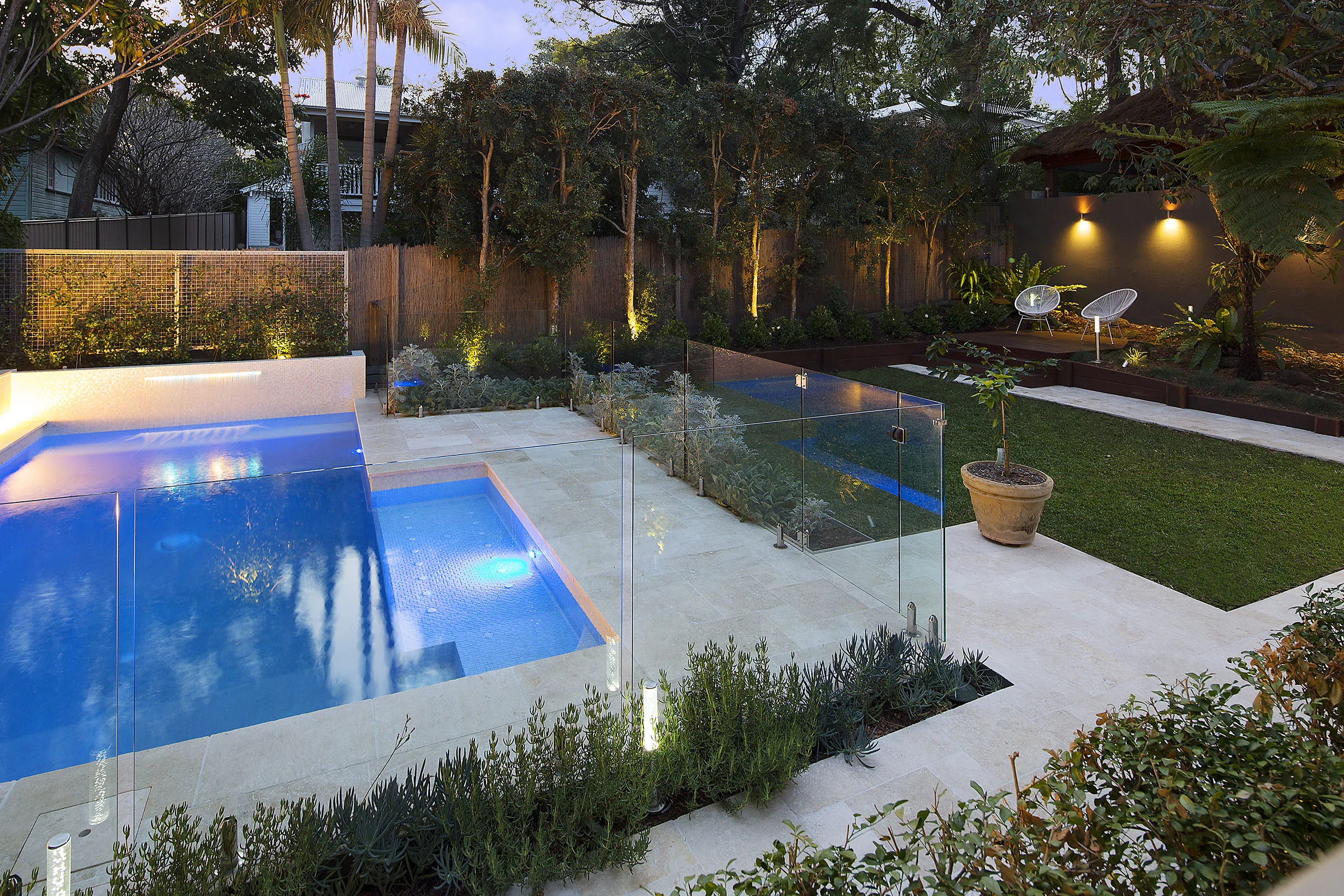 Rectangular pool at night with glass fencing, an uplit hedge screen, and travertine coping
