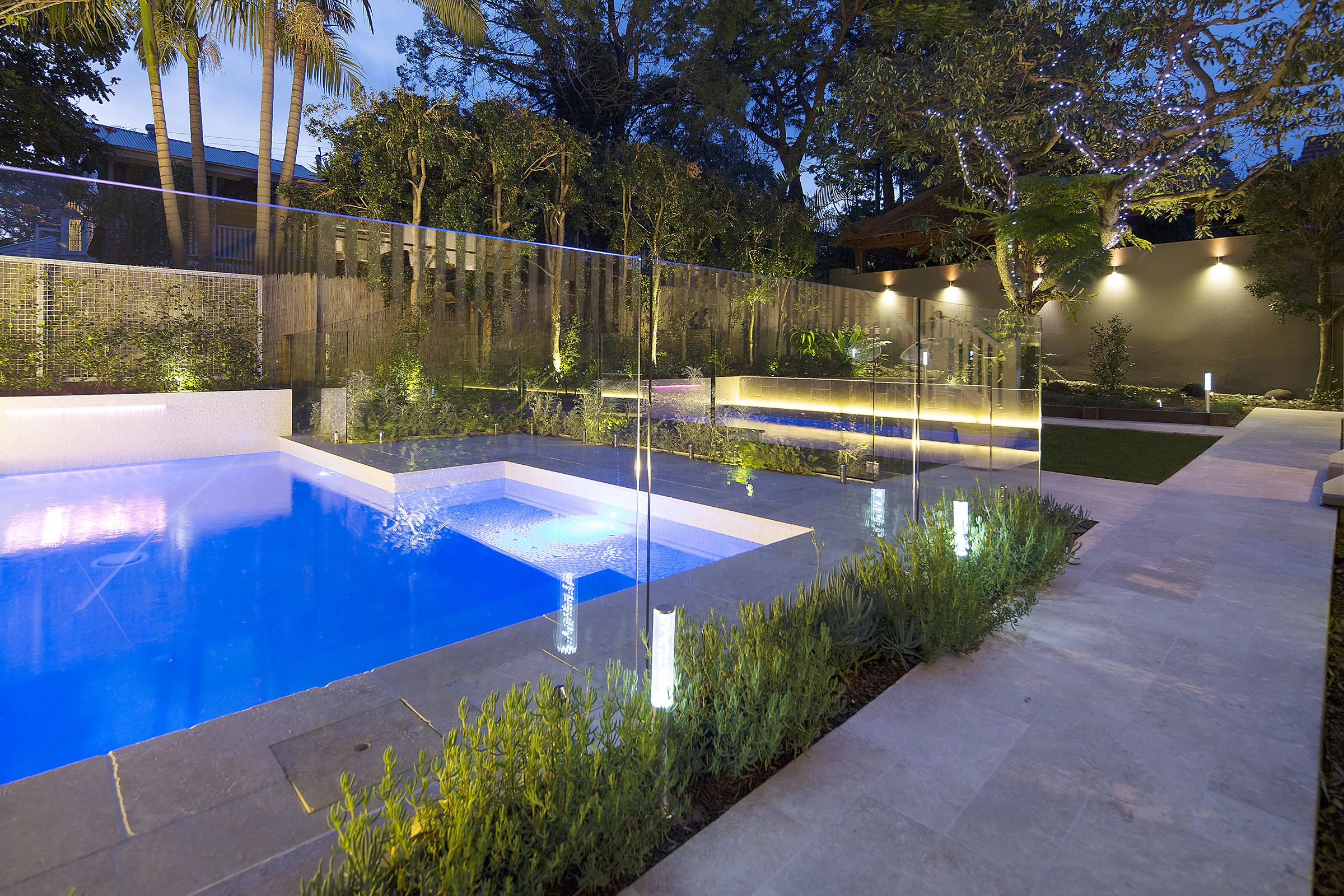 Long rectangular pool at night with glass fencing and travertine paving reflecting the lights