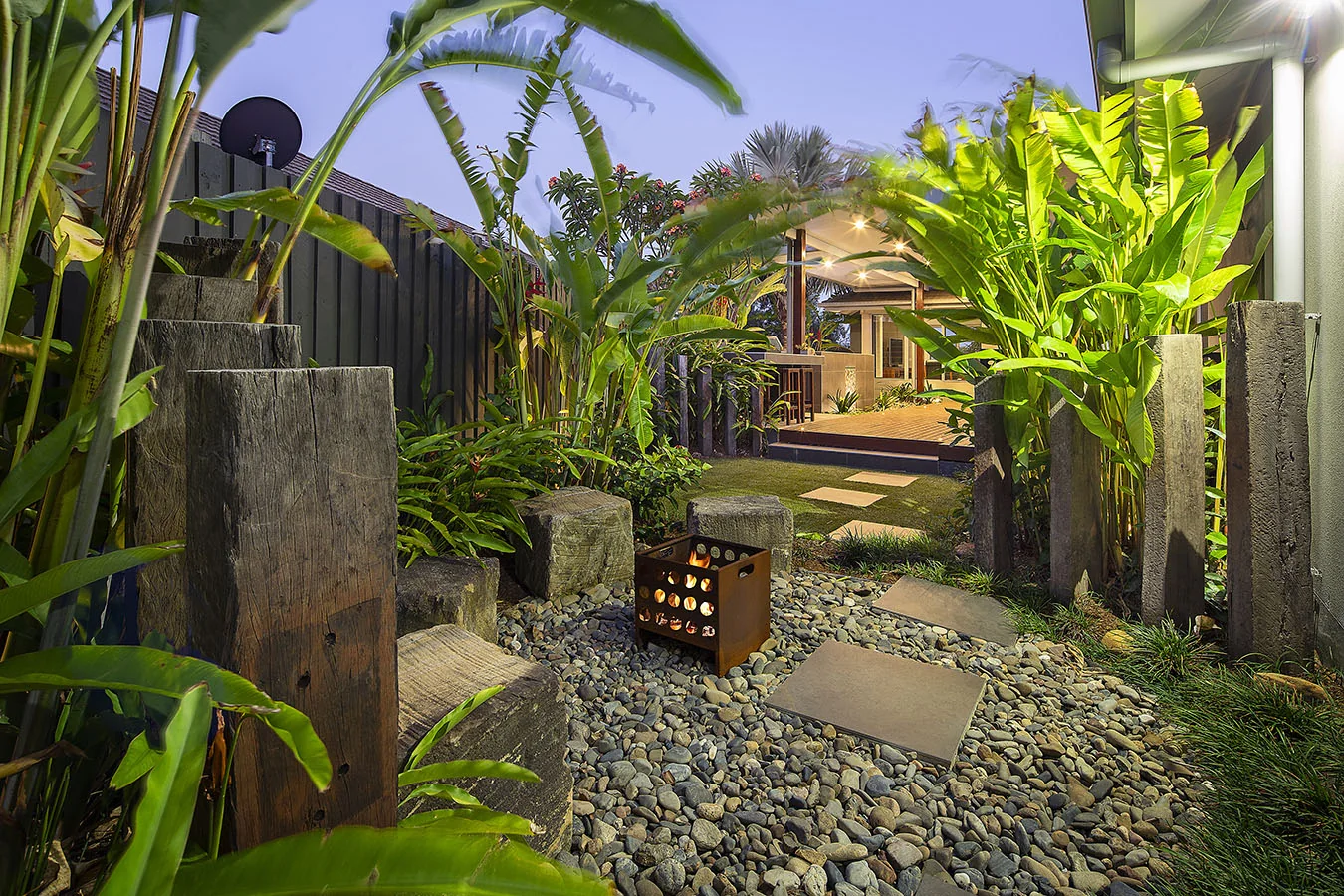 Backlit banana and heliconia foliage framing a stepping path between timber sleeper posts at dusk
