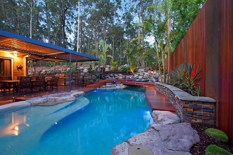 Free-form pool with natural stone edging and a rock waterfall beside a lit timber-lined dining pavilion at dusk