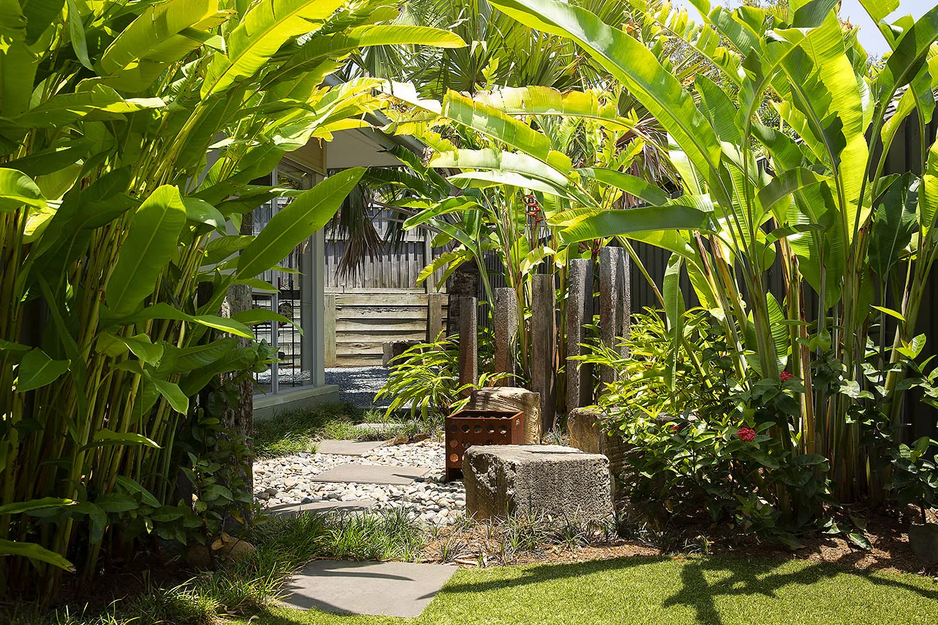 Path through tall golden cane palms with a corten steel stool and dappled light